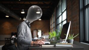 Een persoon met een gloeilamp als hoofd werkt op een computer in een modern kantoor, wat creativiteit en innovatieve ideeën symboliseert.